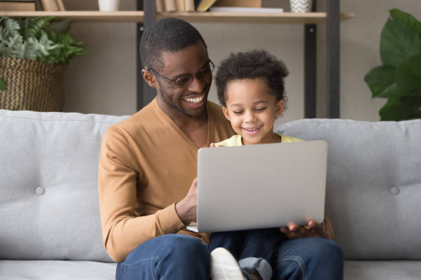 Smiling black young dad sit on couch holding cute little son watching video on laptop together, happy african American father have fun playing computer game with preschooler child relaxing at home