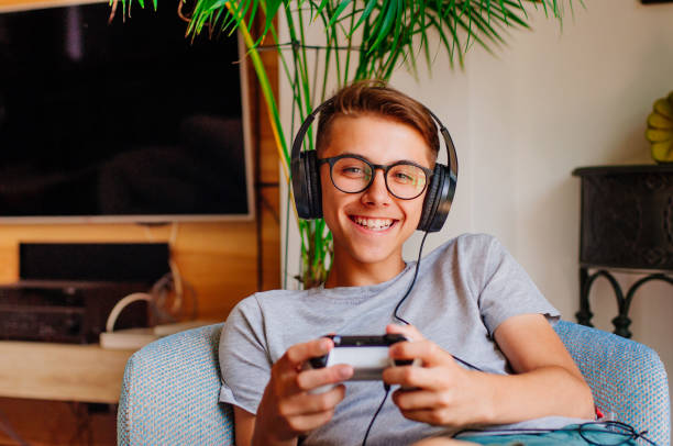 Smiling teenager boy playing console with headphones sitting in the chair indoor looking at camera