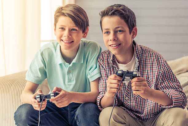 Two attractive teenage boys are playing game console and smiling while sitting on the couch at home